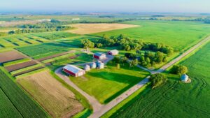 Aerial view of a large, green rural farm with red barns, silos, various fields, trees, and surrounding farmland under a clear blue sky.