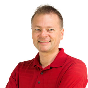 A smiling man with short hair wearing a red polo shirt stands with arms crossed against a plain white background.